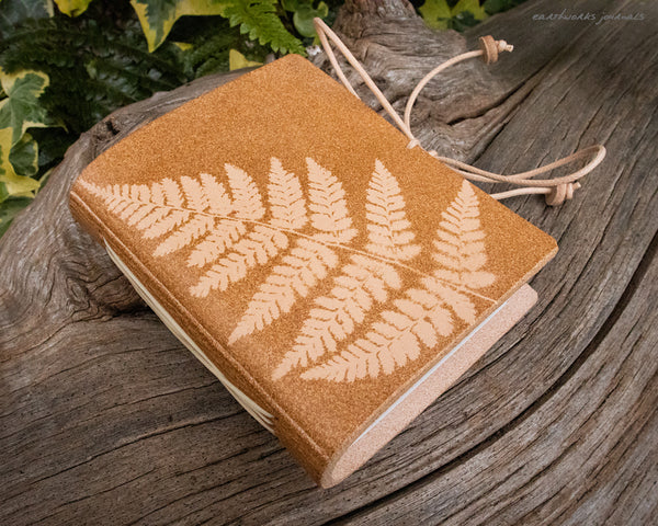 a small pale brown leather journal with a fern leaf design is on weathered wood, made by earthworks journals