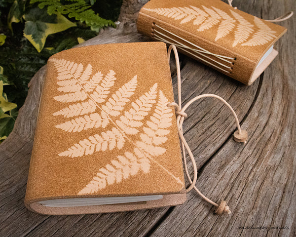 two small pale brown leather journals with fern leaf designs are on weathered wood, made by earthworks journals