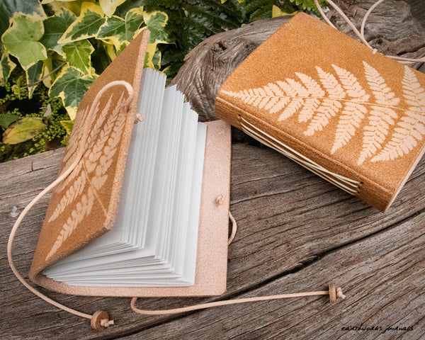 two small pale brown leather journals with fern leaf designs are on weathered wood, made by earthworks journals