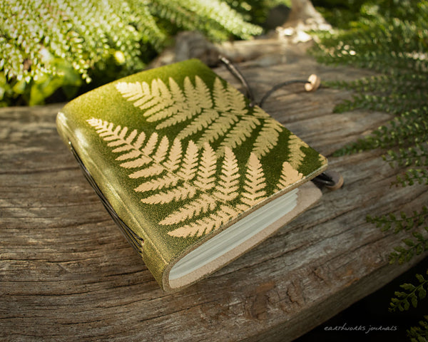 a small green leather journal with a fern leaf design is on weathered wooden planks, made by earthworks journals