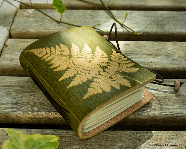 a small green leather journal with a fern leaf design is on weathered wooden planks, made by earthworks journals