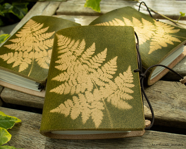 a small green leather journal with a fern leaf design is on weathered wooden planks, made by earthworks journals