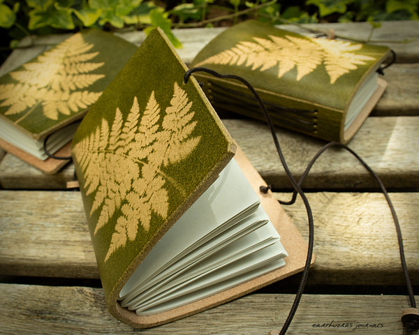 a small green leather journal with a fern leaf design is on weathered wooden planks, made by earthworks journals