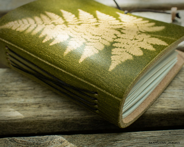a small green leather journal with a fern leaf design is on weathered wooden planks, made by earthworks journals