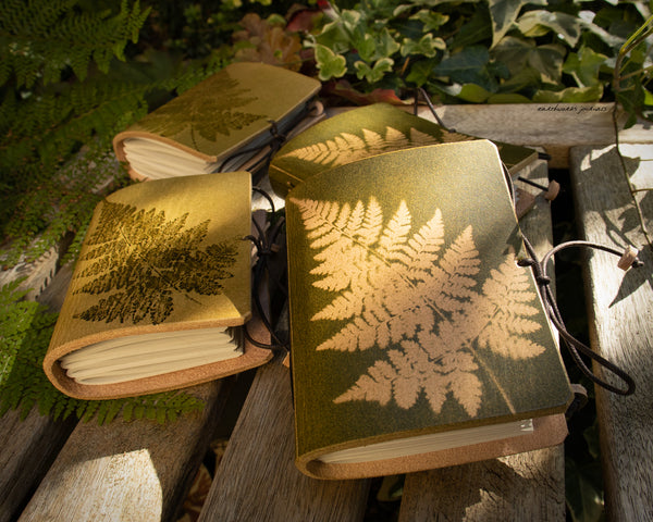 a group of small leather journals with fern leaf designs are on weathered wooden planks, made by earthworks journals