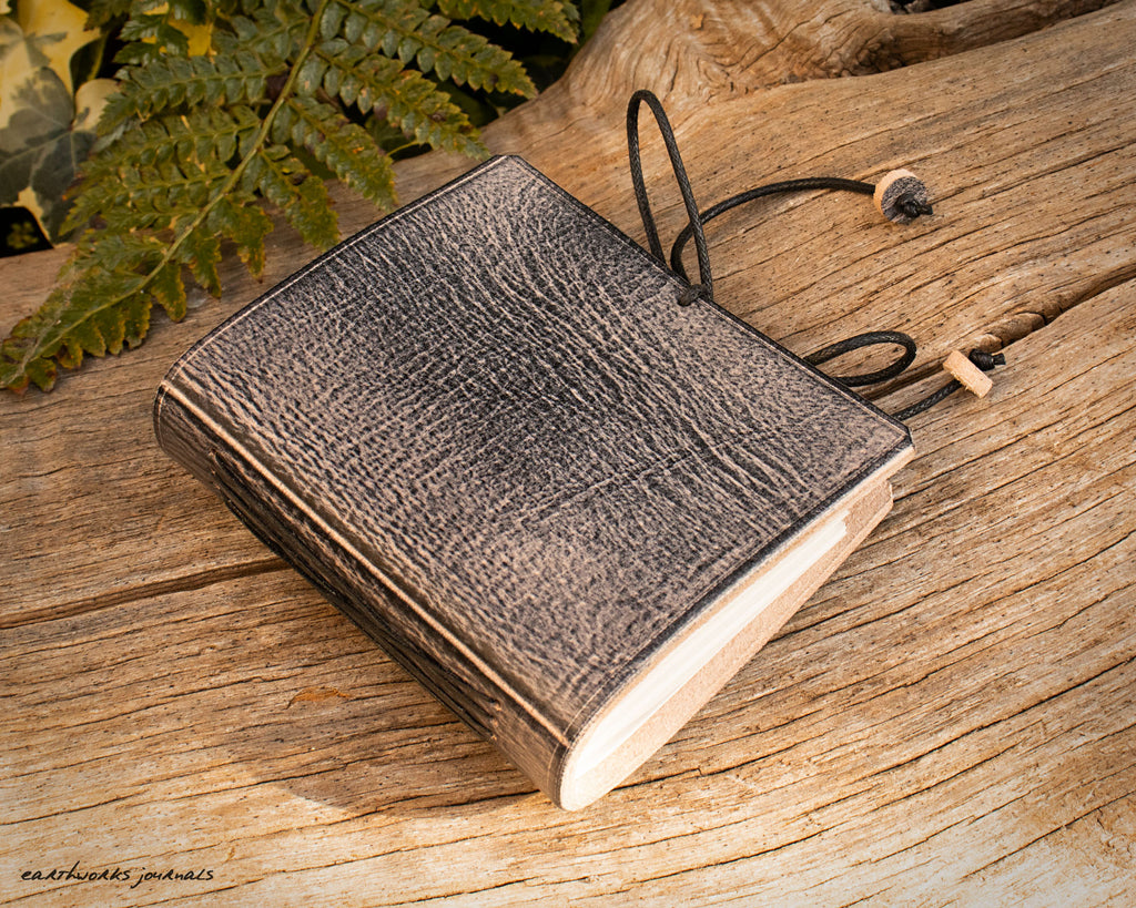 a small grey leather notebook on a piece of weathered wood, made by earthworks journals