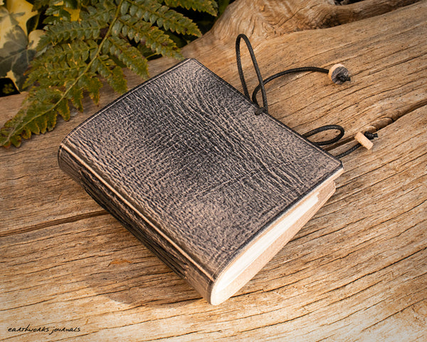 a small grey leather notebook on a piece of weathered wood, made by earthworks journals