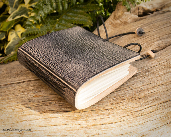 a small grey leather notebook on a piece of weathered wood, made by earthworks journals