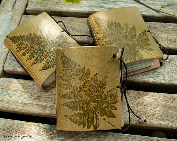three small green leather journals with fern leaf designs are on weathered wooden planks, made by earthworks journals
