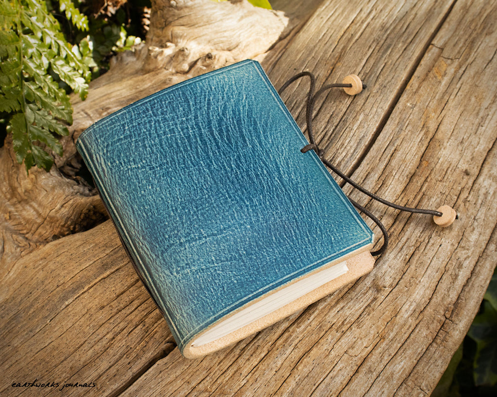 a small blue leather notebook on a piece of weathered wood, made by earthworks journals