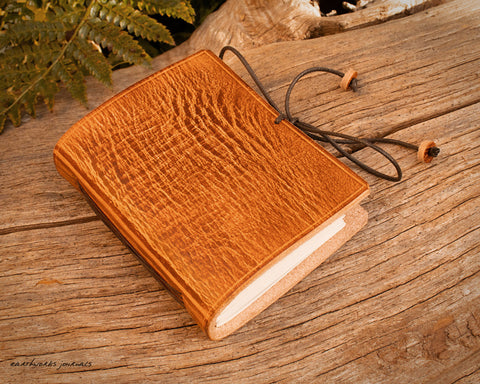 a small brown leather notebook on a piece of weathered wood, made by earthworks journals