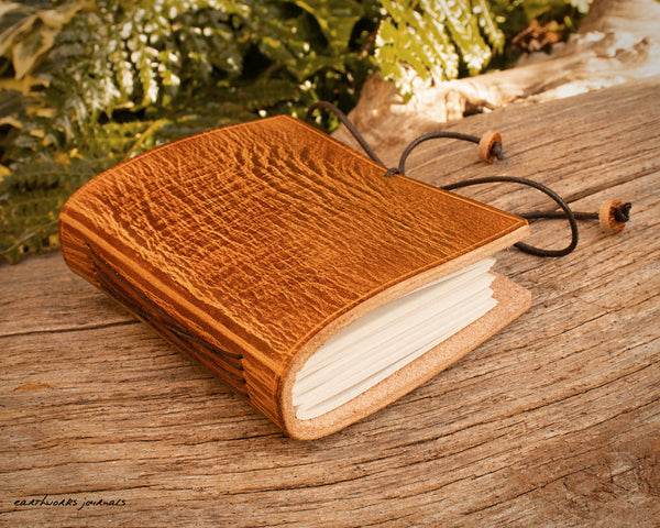 a small brown leather notebook on a piece of weathered wood, made by earthworks journals