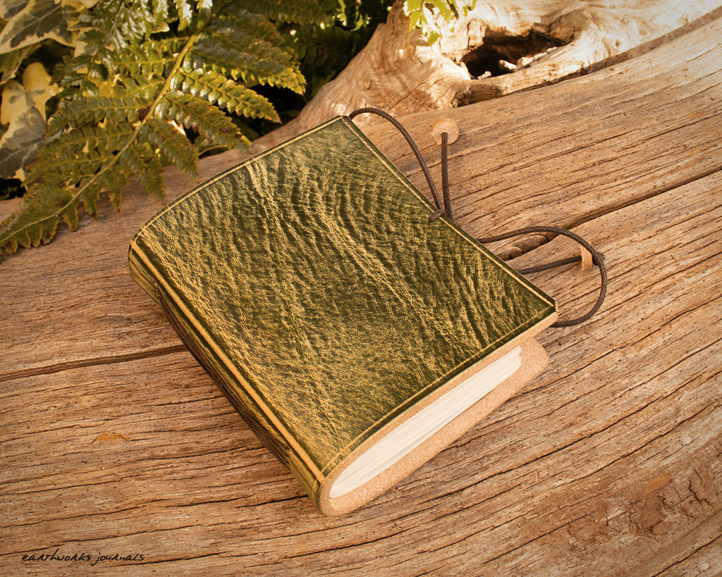 a small green leather notebook on a piece of weathered wood, made by earthworks journals